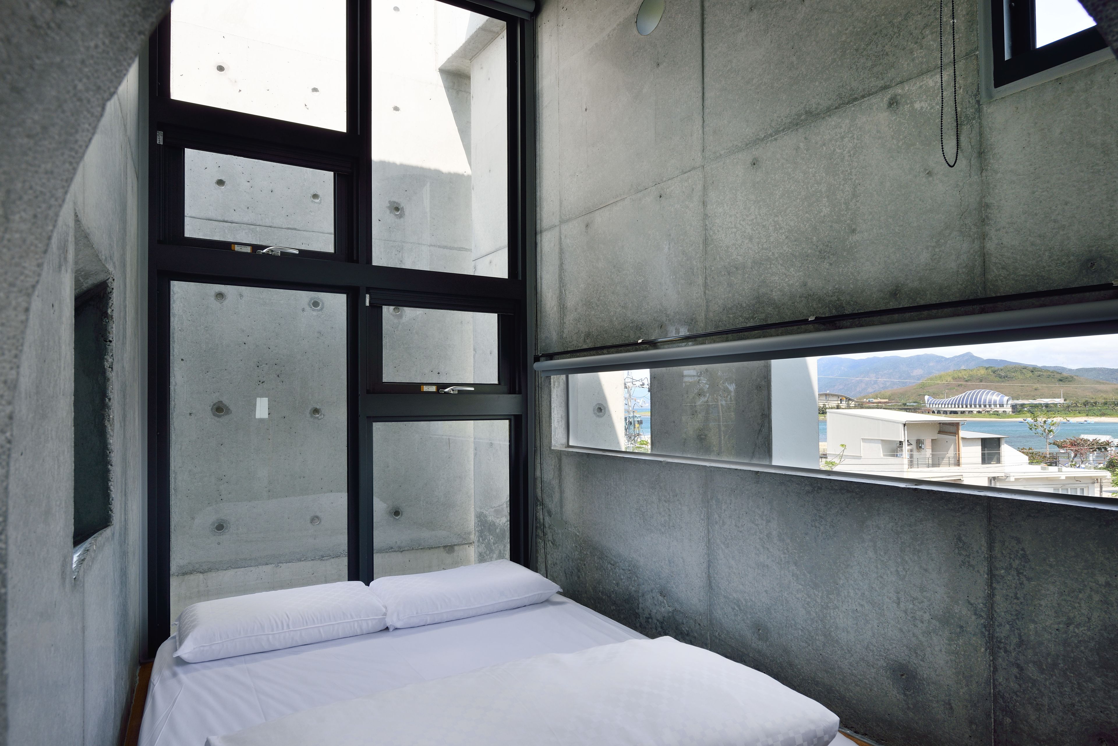 A modern room with a bed, large windows, and a view of buildings and mountains.