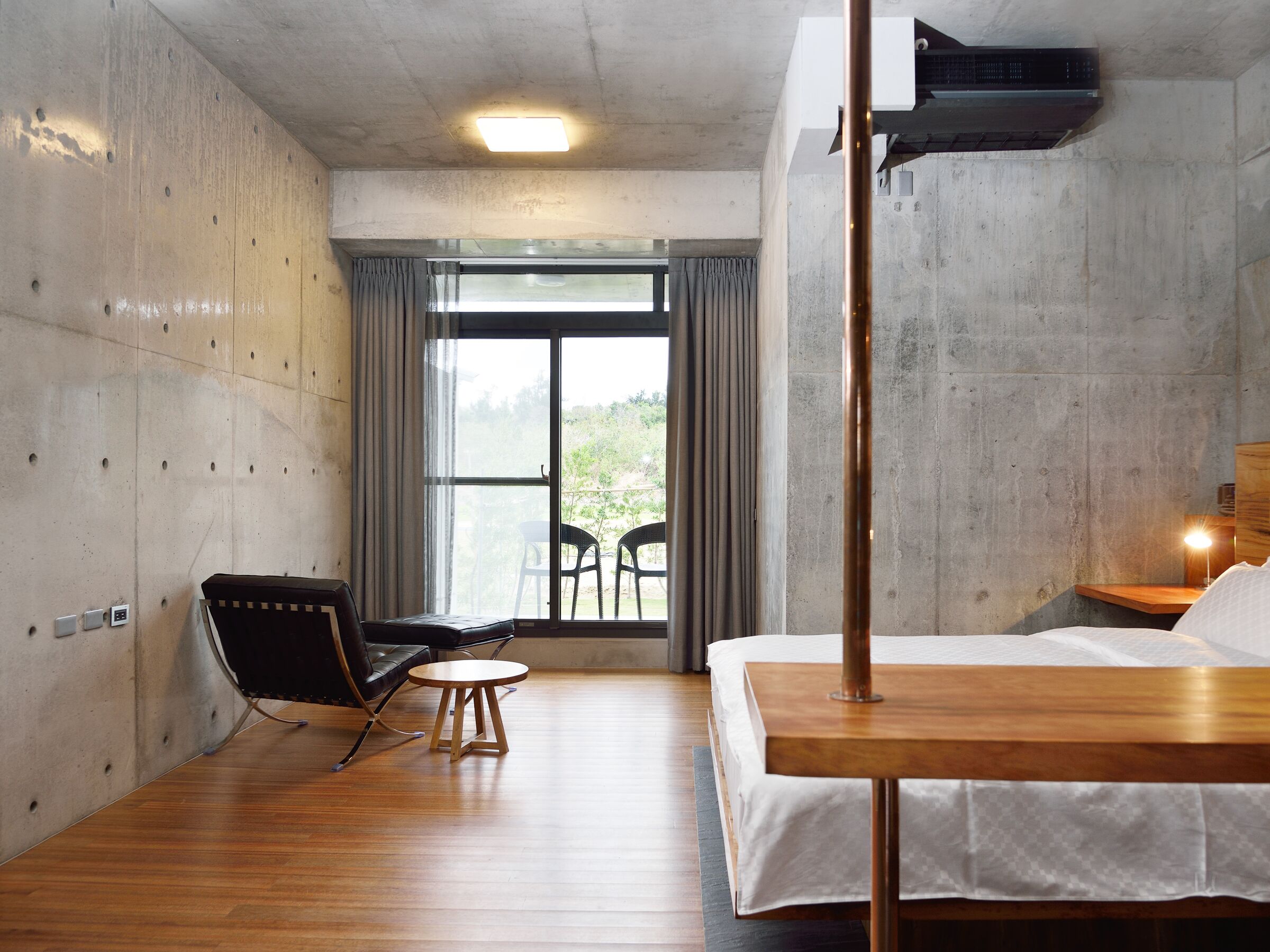 Una habitación de hotel moderna con piso de madera, una cama con colcha blanca, una silla negra, una mesita redonda pequeña y un ventanal grande con cortinas.
