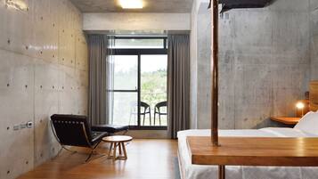 Una habitación de hotel moderna con piso de madera, una cama con colcha blanca, una silla negra, una mesita redonda pequeña y un ventanal grande con cortinas.