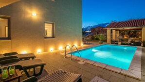 Una piscina con sillas de descanso y zona de barbacoa, rodeada por una casa con iluminación exterior.