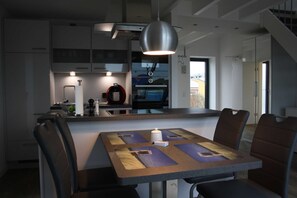 A modern kitchen with a dining area, featuring a large island, overhead lighting, and a view of the outside.