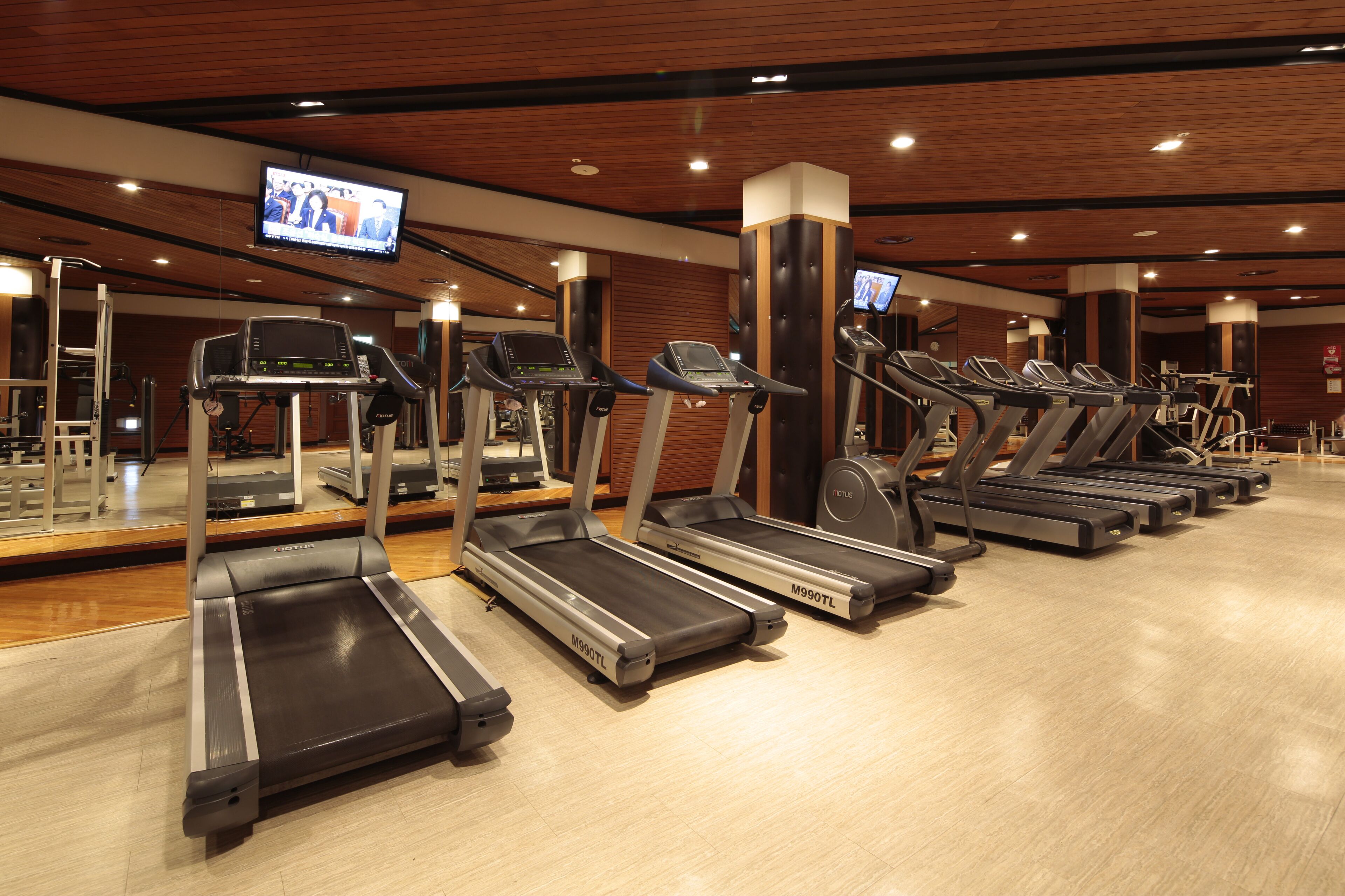 Un gimnasio con varias cintas de correr alineadas en filas, una pantalla de televisión montada en el techo y paneles de madera en las paredes.