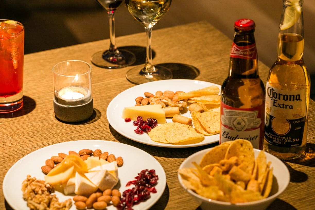 A table with plates of cheese, nuts, and crackers, a bottle of Corona Extra beer, and glasses of wine.