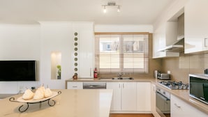 A modern kitchen with white cabinetry, a central island with a dish rack, a built-in oven, and a window with blinds. - BOUTIQUE STAYS - Sandy Breeze 3 (Sandringham)