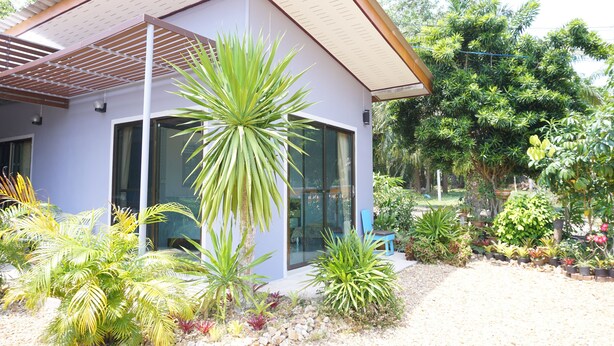 A modern house with a covered patio, surrounded by various tropical plants and a gravel driveway. - Kedsila Hometel (Pathio)