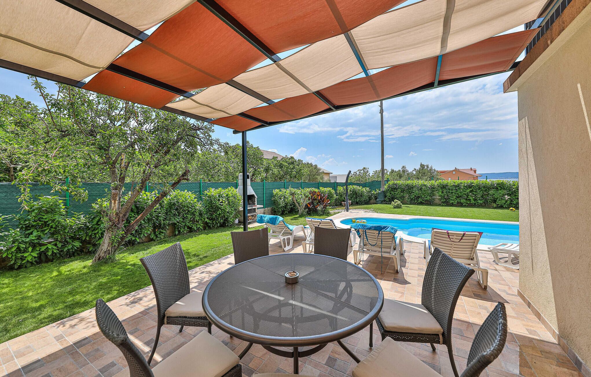 Een terras buiten met een ronde tafel en stoelen, een zwembad op de achtergrond en een overdekt gedeelte met uitzicht op zee.