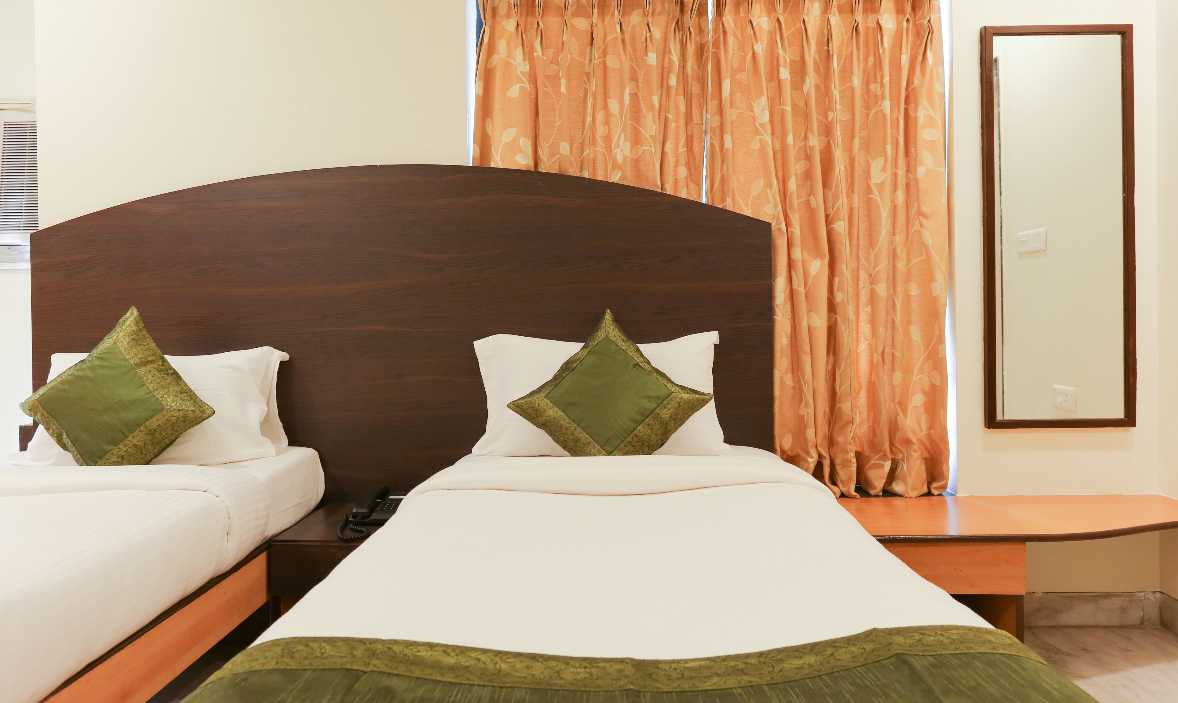 Een hotelkamer met een houten hoofdeinde, twee bedden met witte lakens en groene kussens, een nachtkastje en een spiegel aan de muur.