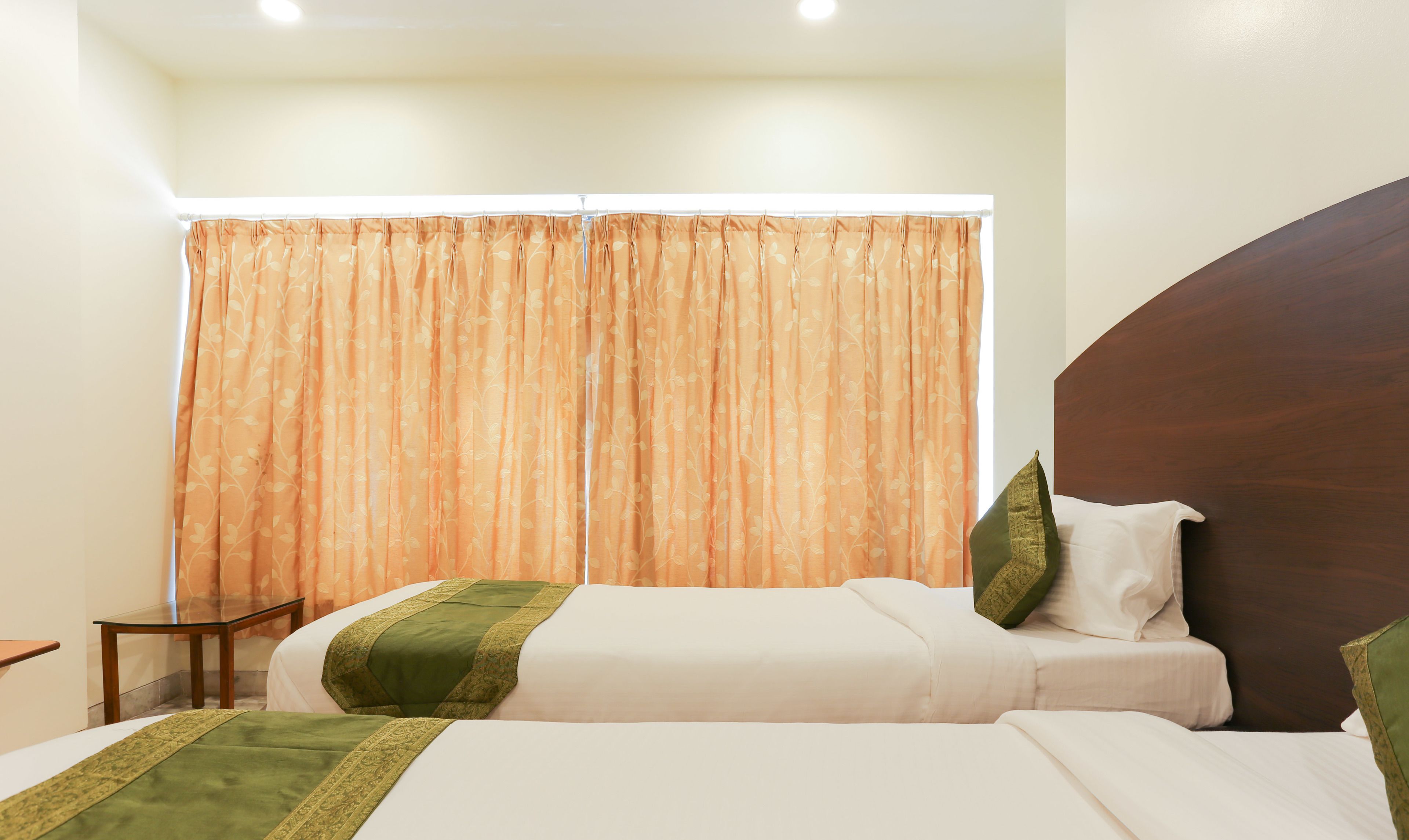 Habitación de hotel con dos camas, cabecero de madera, una mesita y una ventana con cortinas naranjas.