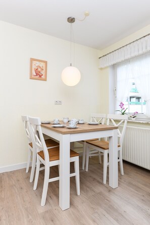 Un coin repas avec une table en bois et des chaises blanches, un luminaire suspendu et une fenêtre avec des rideaux.