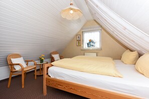 Une chambre avec un plafond en pente, un lit recouvert de linge de lit blanc, deux chaises en bois, une petite table ornée de fleurs et une fenêtre donnant sur un immeuble.