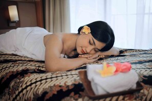 A person lying on a bed with a patterned bedspread, a pillow, and a tray with a towel and flowers.