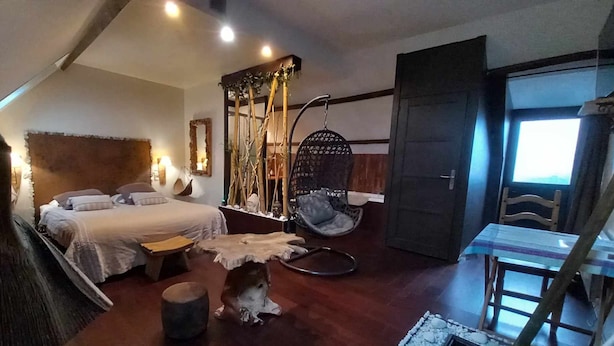Un dormitorio con cama, una silla colgante, una mesita y una puerta que da a otra habitación.