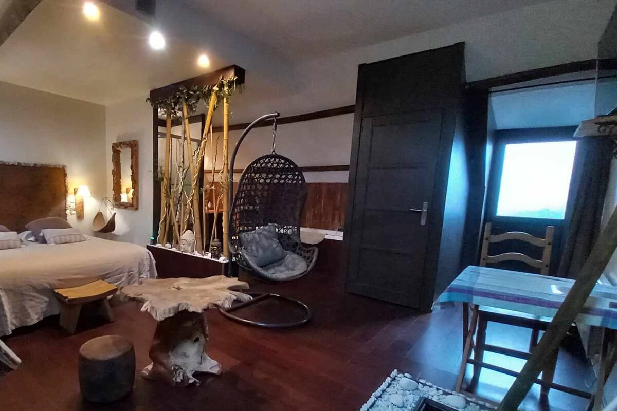 Une chambre à coucher avec un lit, un fauteuil suspendu, une petite table et une porte donnant sur une autre pièce.