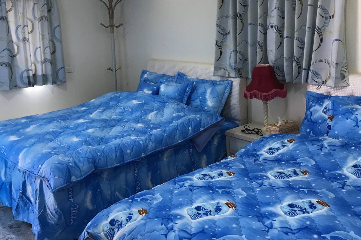 A bedroom with two beds, a red lamp, and a window with patterned curtains.