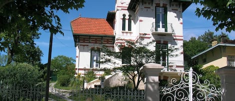 Un edificio blanco con techo rojo y detalles verdes, rodeado de árboles y una cerca de hierro forjado blanco.