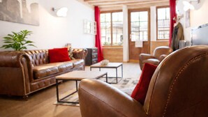 Una sala de estar con un sofá de cuero marrón, una mesa de centro y un cojín rojo. Hay una planta en la esquina y una ventana con cortinas.
