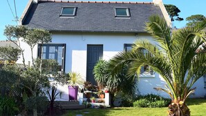Une maison à deux étages avec un toit recouvert de tuiles grises, des murs blancs et une porte bleu foncé. Il y a deux fenêtres à l’étage supérieur et un petit porche agrémenté de plantes en pot et d’un palmier.