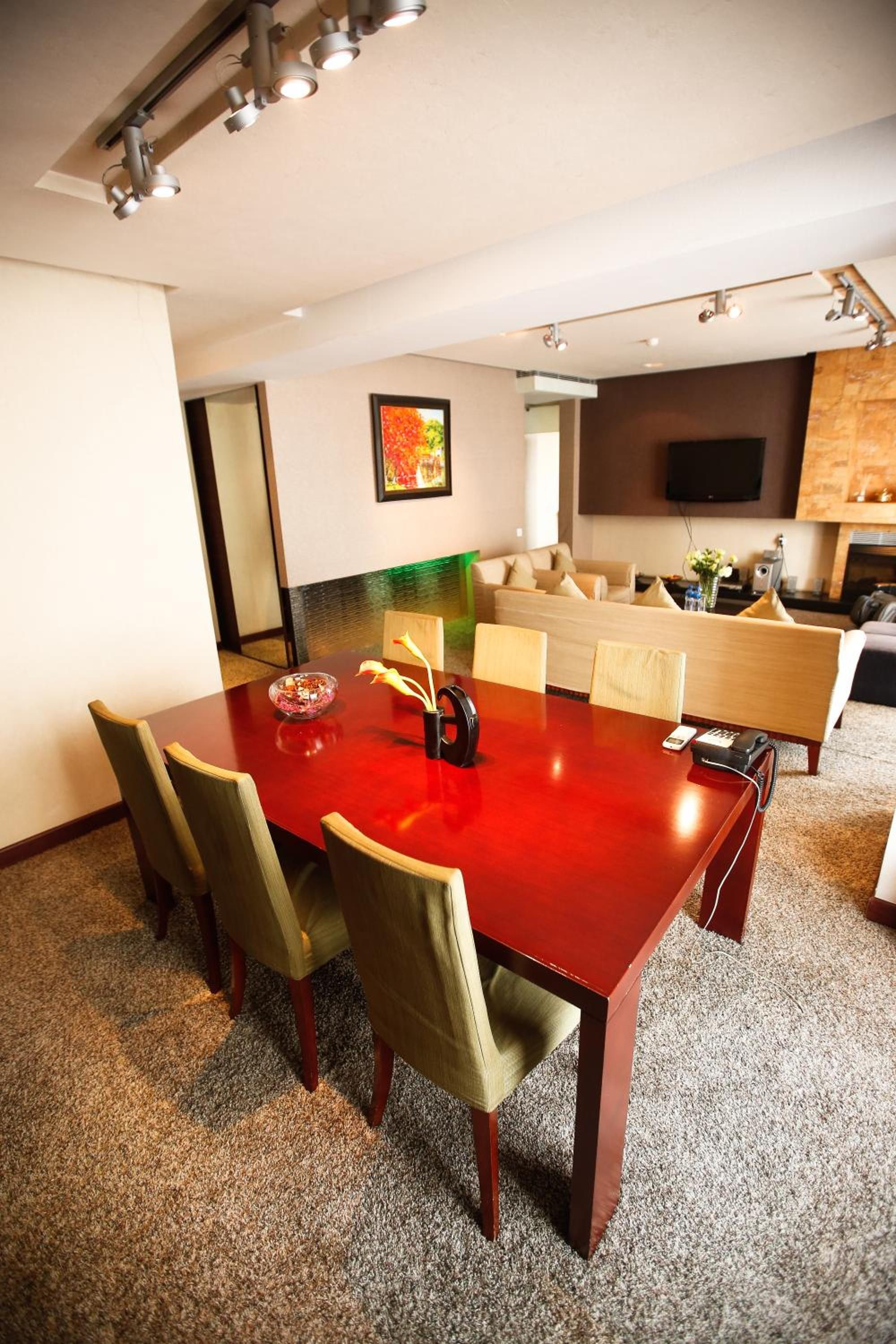 Un salón de conferencias con una mesa roja, sillas verdes, un jarrón con flores y un cuadro en la pared.