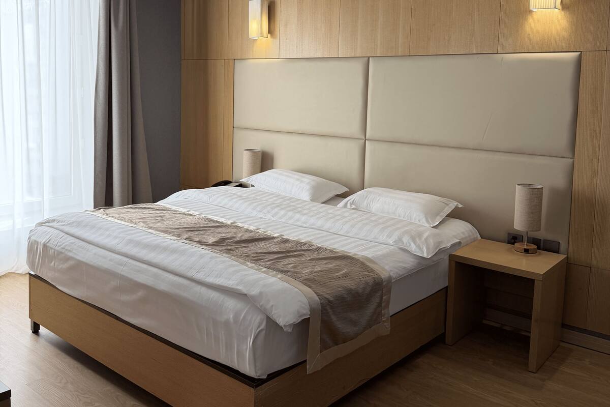 Moderni hotellihuone, jossa on suuri sänky, puinen pääty, yöpöytävalaisimet ja näkymä ulos.