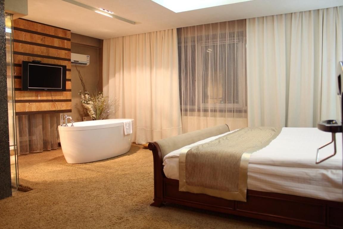 A hotel room with a large bed, a bathtub, a television mounted on a wooden panel, and a window with sheer curtains.