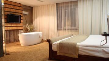 A hotel room with a large bed, a bathtub, a television mounted on a wooden panel, and a window with sheer curtains.