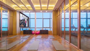 Un gimnasio espacioso con suelos de madera, colchonetas de ejercicio, una pantalla de televisión y ventanas amplias.