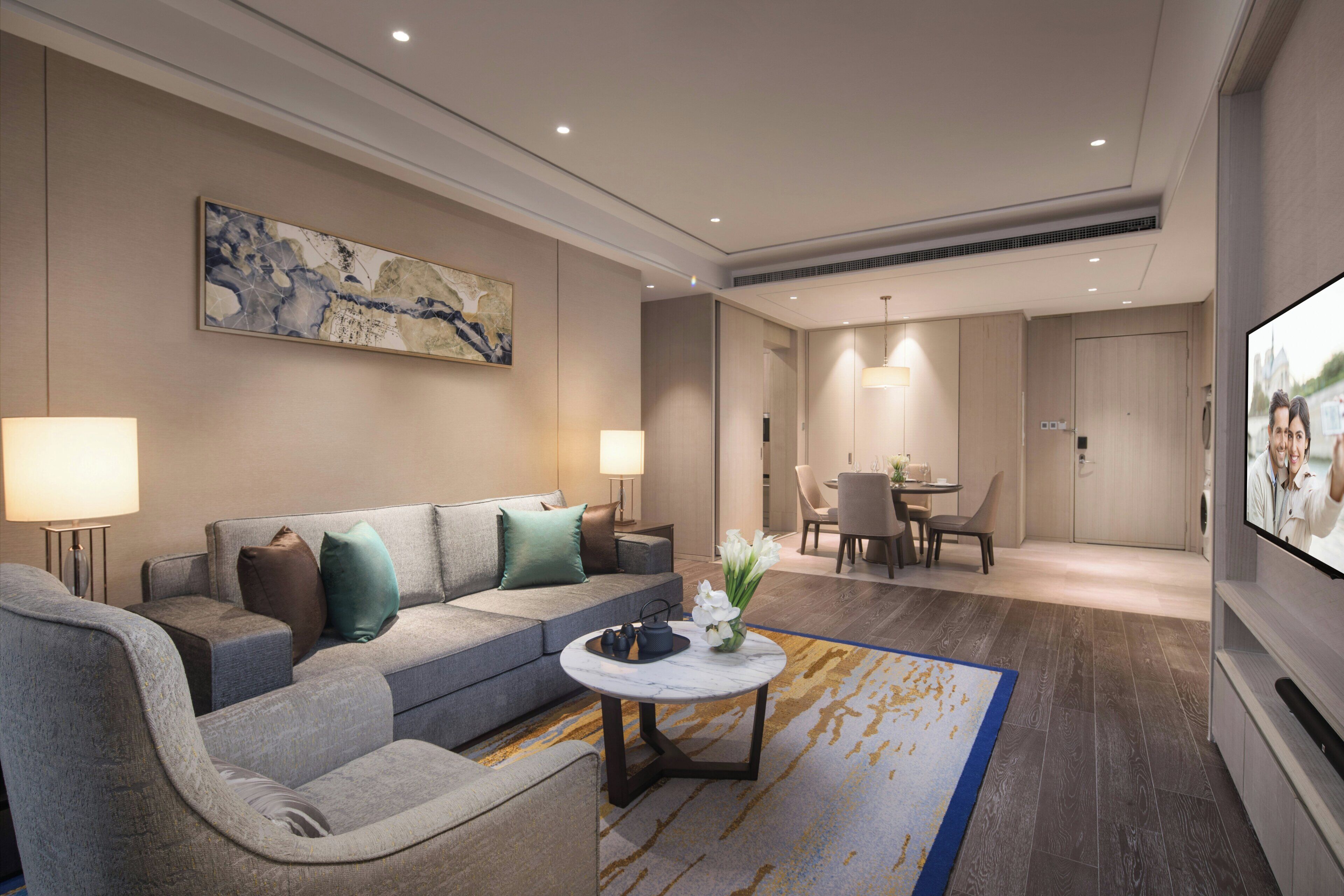 A modern hotel room with a grey sofa, a round coffee table, and a dining area with a table and chairs.