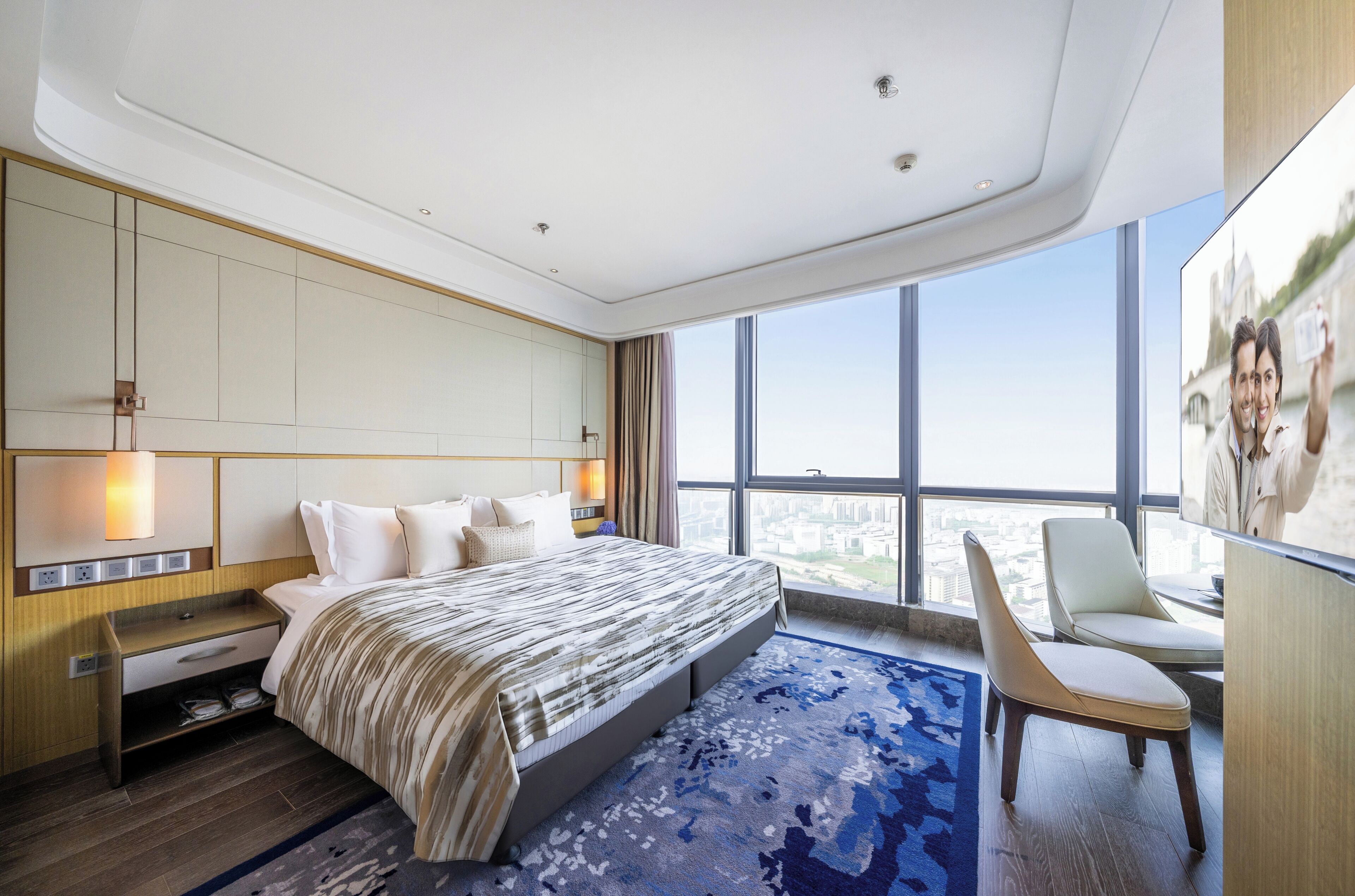 A modern hotel room with a large bed, a desk, a chair, and a view of the city through large windows.