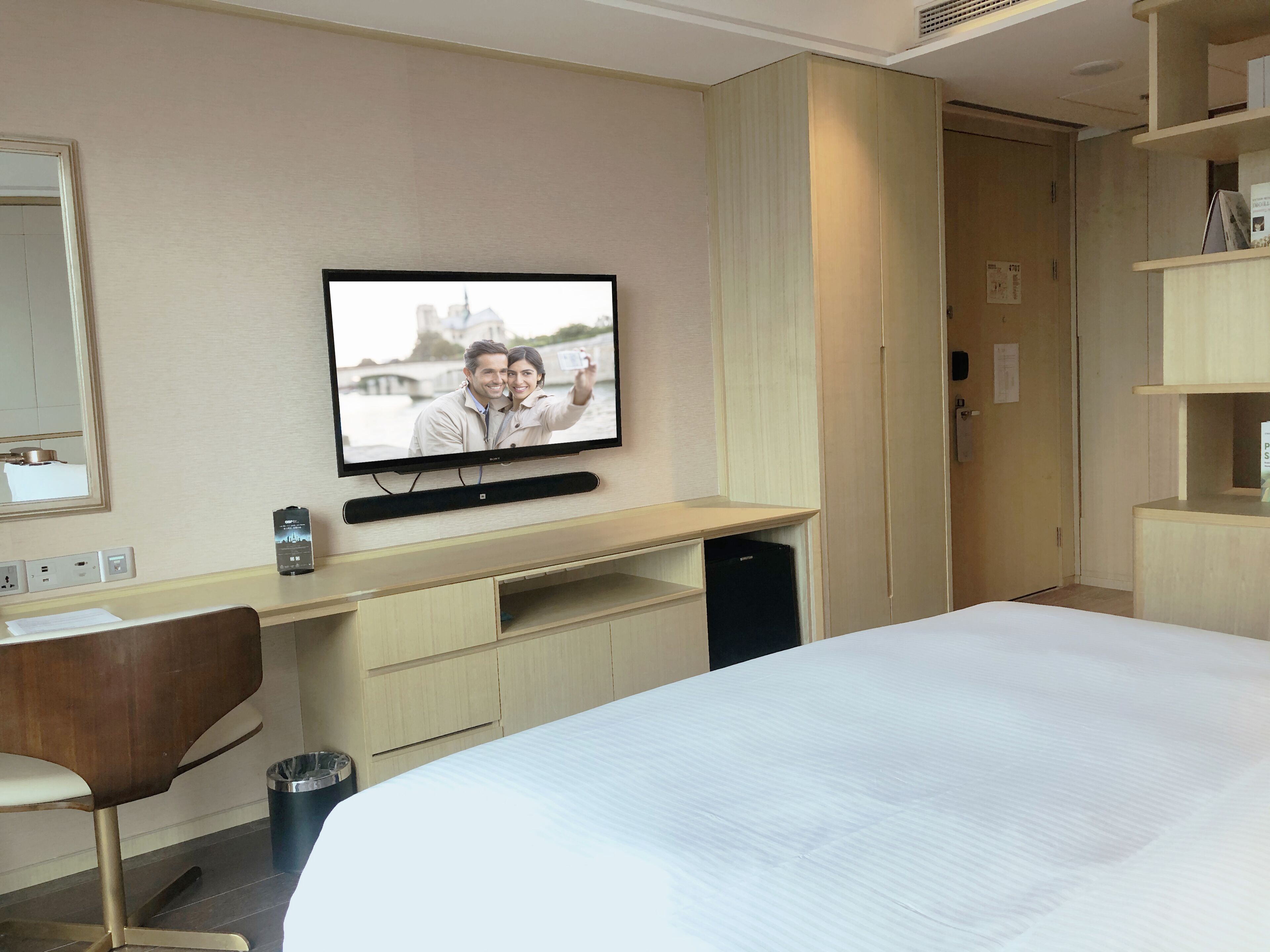 Una habitación de hotel con cama, escritorio con computadora, silla, televisor montado en la pared y un cesto de basura.