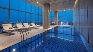 Una piscina interior moderna con baldosas azules cristalinas, sillones de descanso y vistas a la ciudad a través de amplios ventanales.
