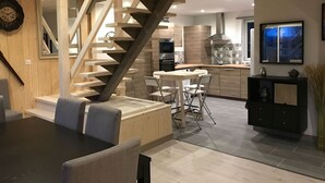 Une cuisine moderne dotée d’un îlot central, d’un coin repas avec une table noire et des chaises grises, et d’un escalier en bois menant à un étage supérieur.