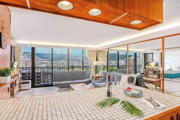 A spacious living room with a large window, a sofa, a coffee table, and a dining area with a table set for two.