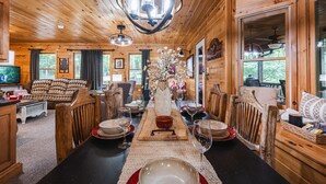 Interior de una cabaña de madera con comedor listo para una comida, mostrando una mesa larga, sillas y una araña.
