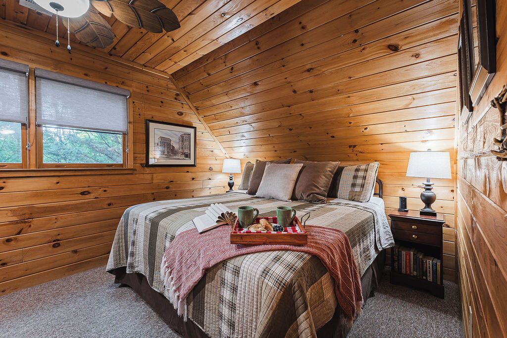 Une chambre confortable aménagée dans une cabane en bois, avec un lit, deux lampes de chevet, une table de chevet avec des livres, une fenêtre donnant sur un paysage agréable et un tableau encadré au mur.