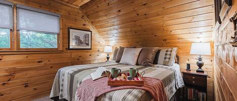 Une chambre confortable aménagée dans une cabane en bois, avec un lit, deux lampes de chevet, une table de chevet avec des livres, une fenêtre donnant sur un paysage agréable et un tableau encadré au mur.