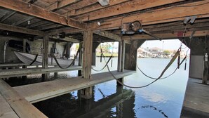 Un muelle de madera con un barco atracado a la izquierda, una hamaca colgada del techo, y vista al agua y edificios al fondo.