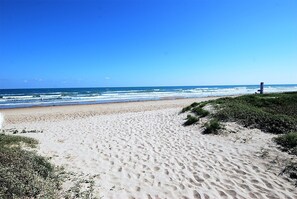 Una playa arenosa con huellas que conducen al agua, un cielo despejado y una estructura a lo lejos.
