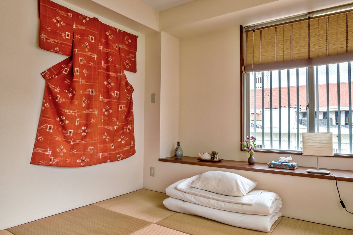 Een kamer met tatami vloeren, een futonbed, een raam met bamboeblamen en een rood gordijn met een patroon.