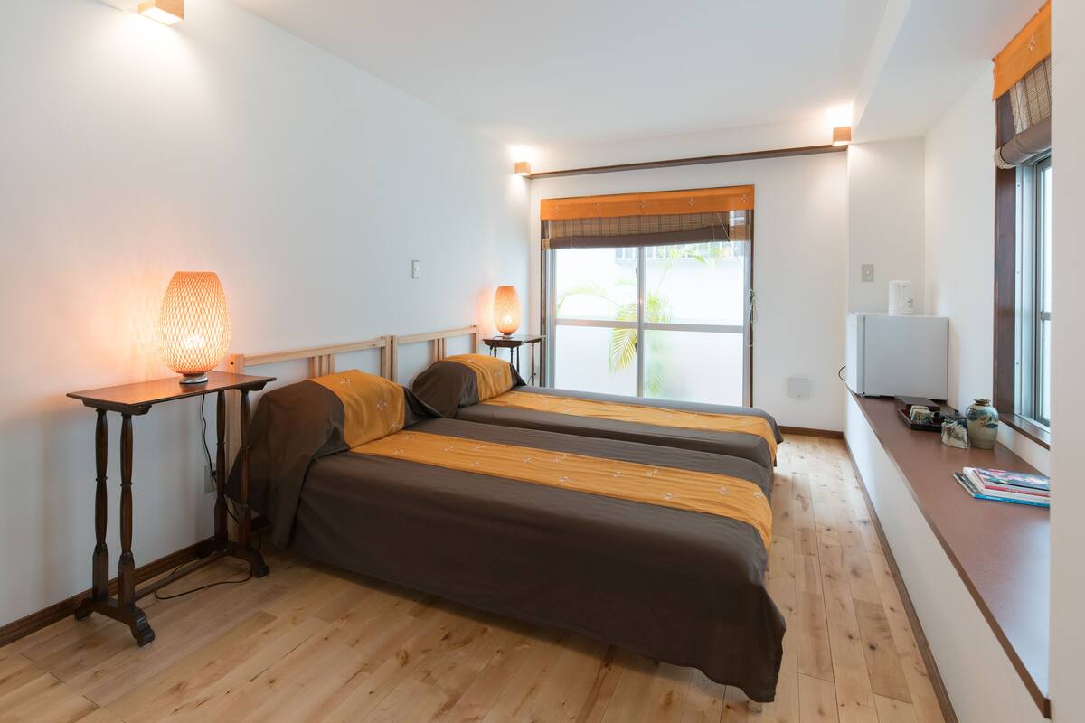 Een kamer met twee bedden, een houten vloer, een bureau met lamp, een raam met jaloezieën en een koelkast.