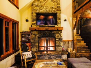 A cozy living room with a stone fireplace, a television mounted above it, and a wooden staircase leading to an upper level.