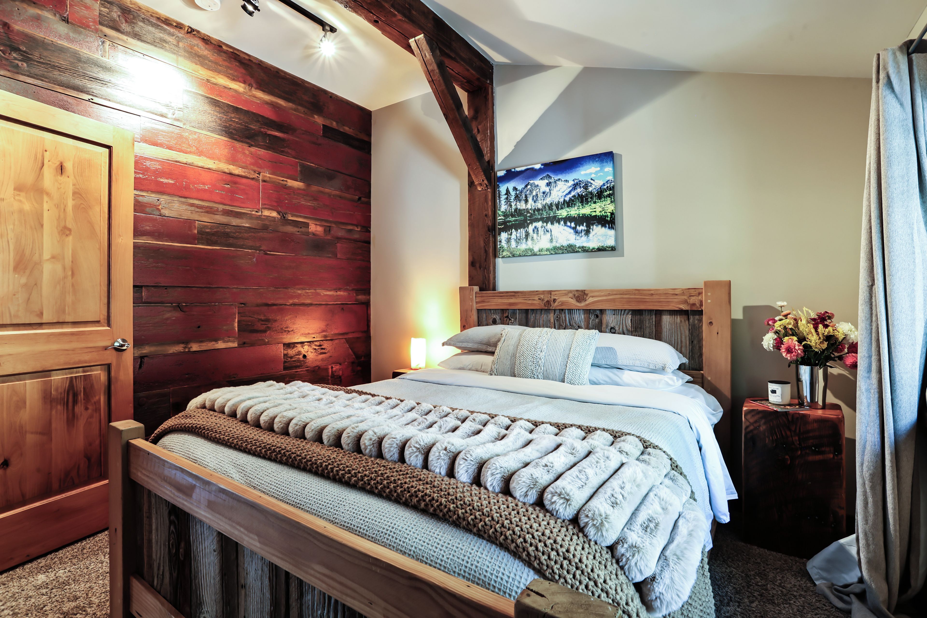 A bedroom with a wooden headboard, a bed with a white and brown blanket, a nightstand with a lamp, and a painting of a mountain landscape on the wall.