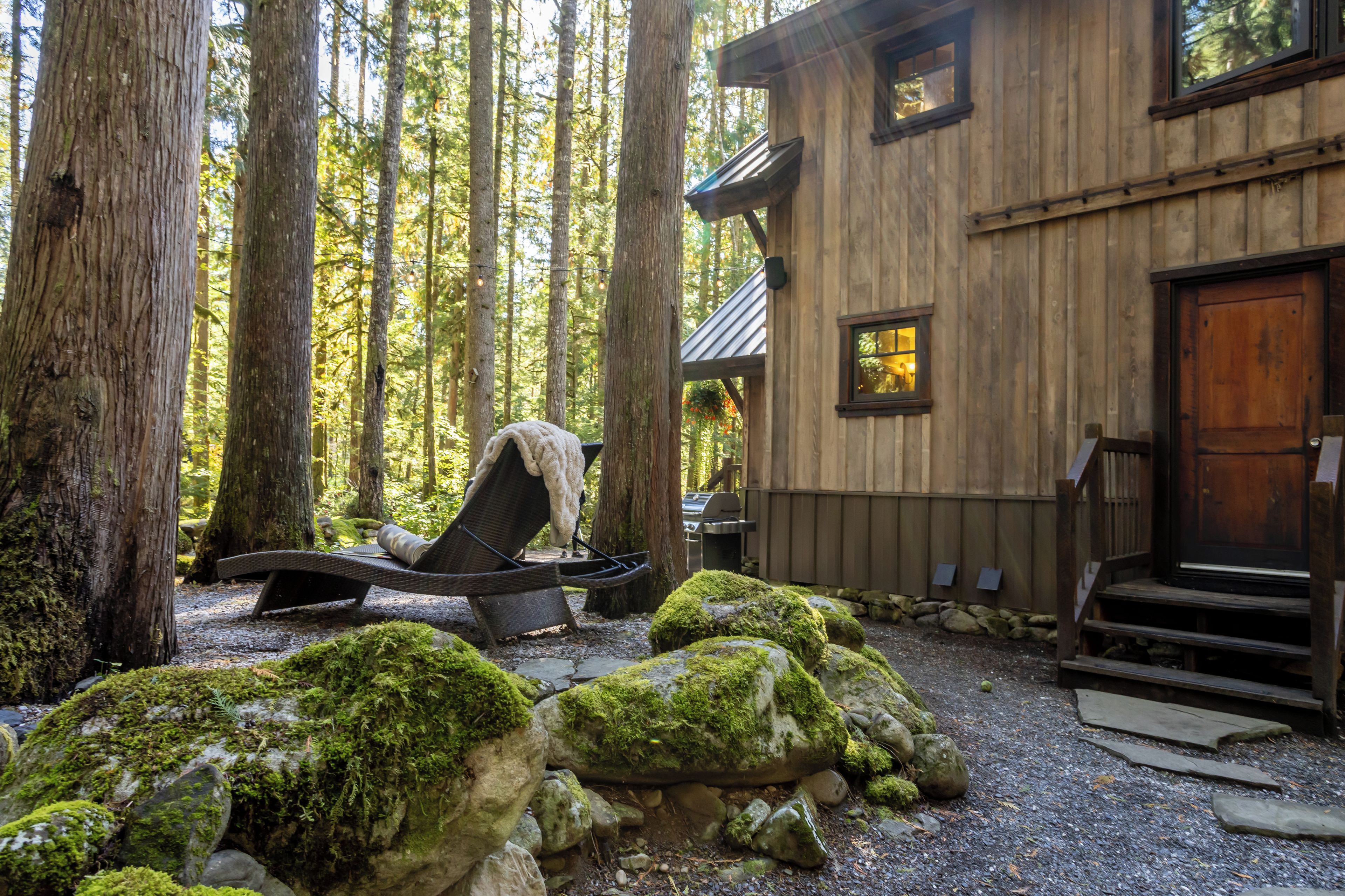 A rustic cabin with a covered outdoor seating area, surrounded by large trees and moss-covered rocks.