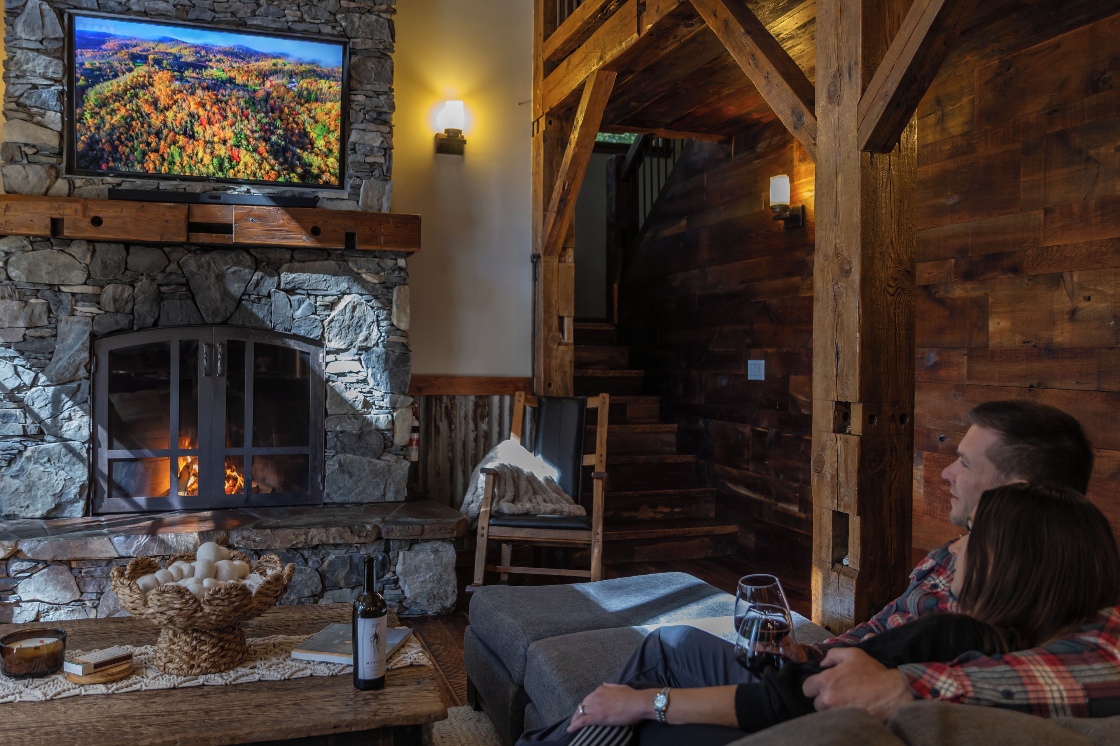A cozy cabin interior with a stone fireplace, a television mounted on the wall, and a wooden staircase.