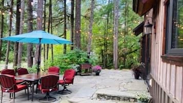 An outdoor patio with a stone table and red chairs, surrounded by tall trees and string lights.