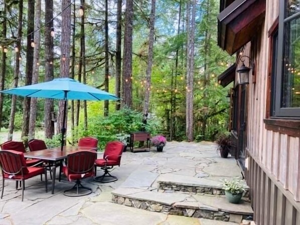 An outdoor patio with a stone table and red chairs, surrounded by tall trees and string lights. - Knotty Lodge – Rustic Charm + Modern Comfort near Mt. Baker, Hot Tub, WiFi (GLACIER)