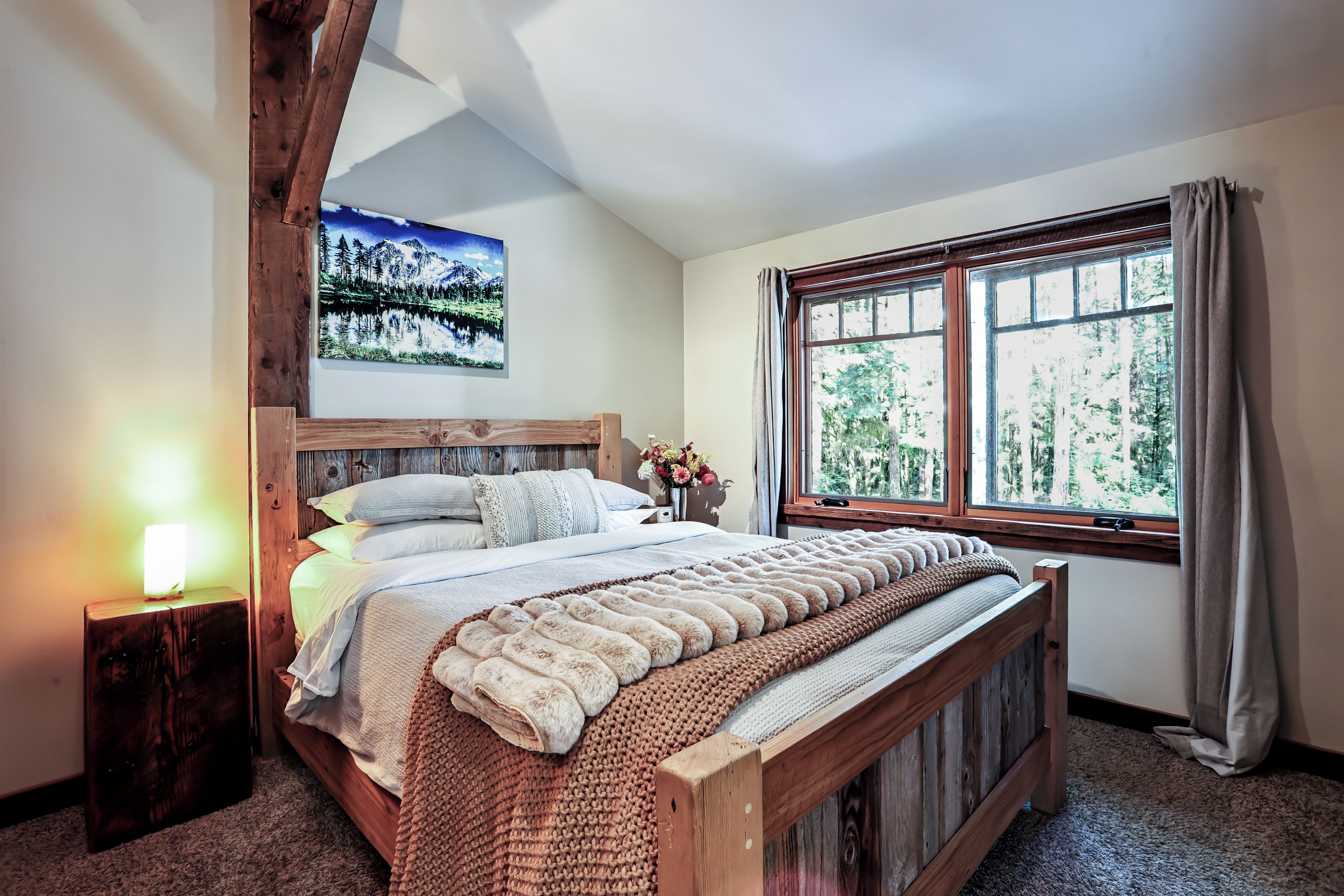 Una camera da letto con un letto in legno, un comodino, una lampada a parete, una finestra con tende e un quadro incorniciato appeso al muro.