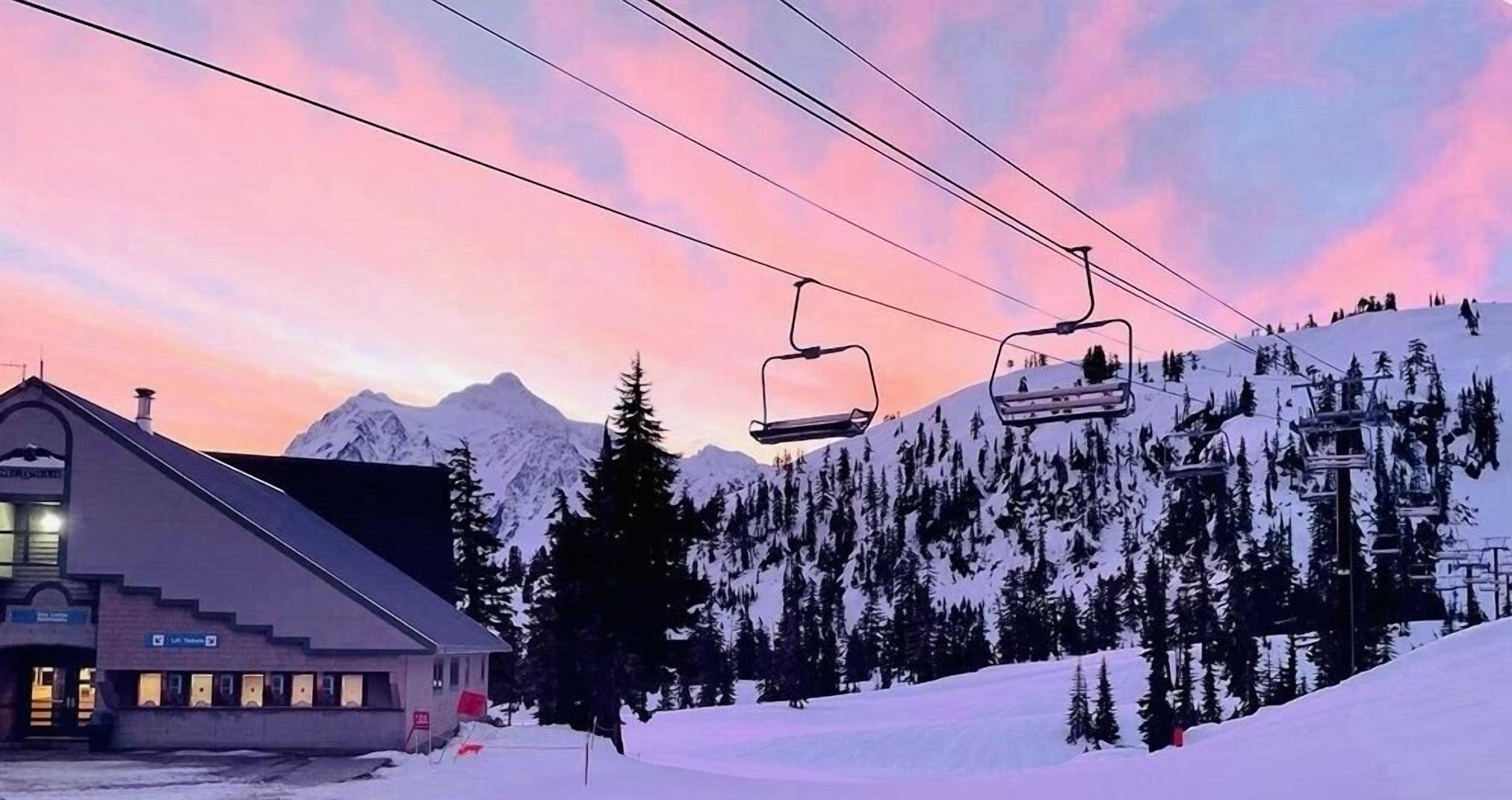 A ski lift chair in motion against a backdrop of snow-covered mountains and a building with a sloped roof.