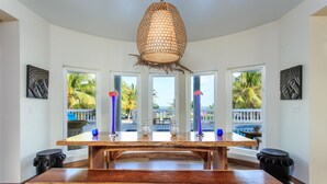Een ruime eethoek met een grote houten tafel, een hangende geweven lamp en grote ramen met uitzicht op palmbomen en een zwembad.