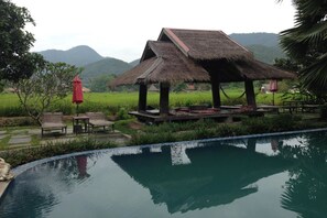 A poolside area with a thatched-roof gazebo, lounge chairs, and a view of a green field and mountains in the background. - Pai Chan Cottage & Cuisine (Pai)