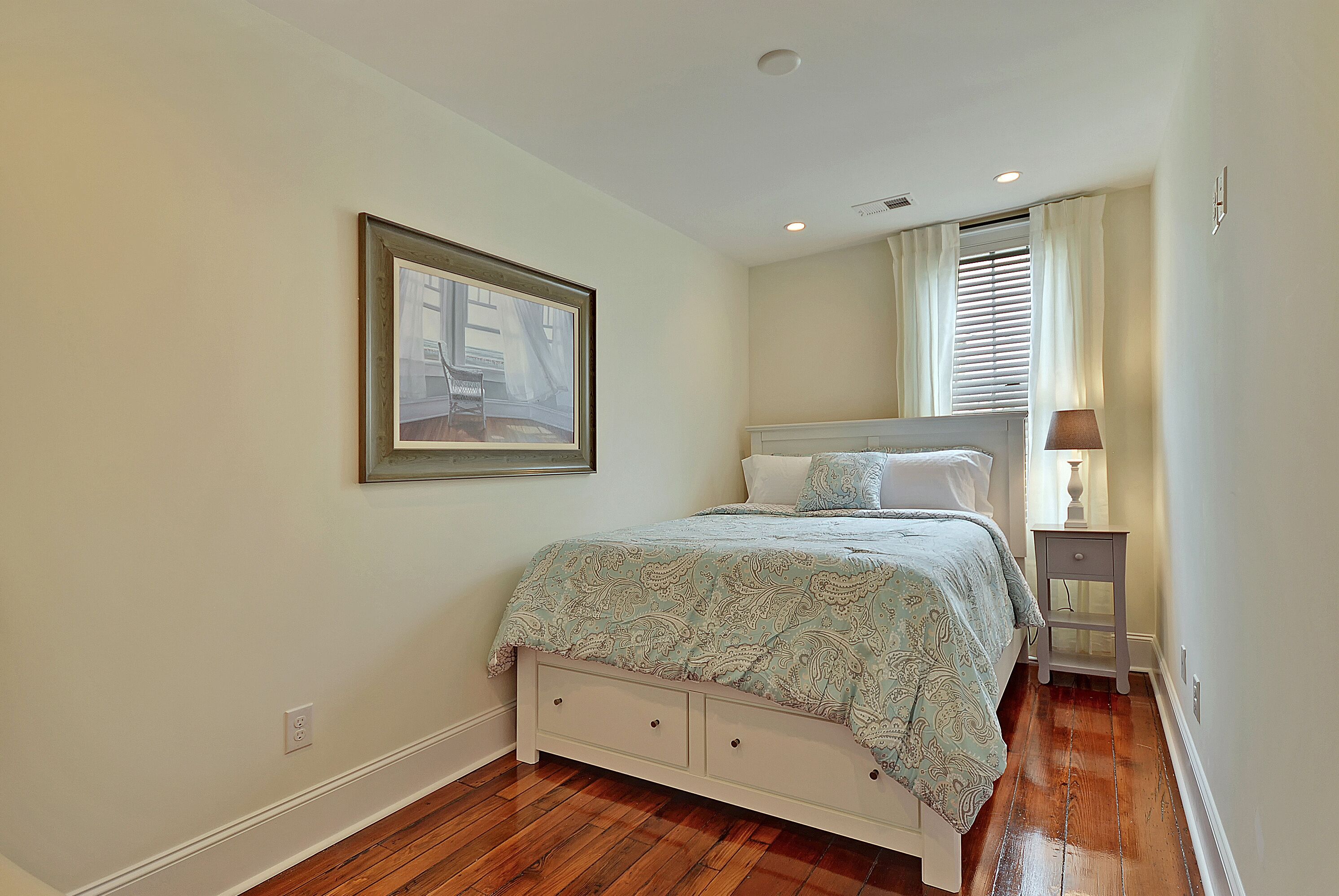 A bedroom with a bed, a nightstand, a lamp, a framed picture on the wall, and a window with curtains.